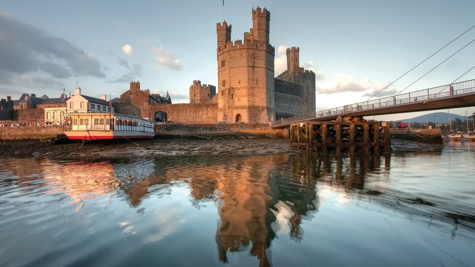 Banded sandstone curtain walls and polygonal towers of Caernarfon Castle rising above the harbour at Caernarfon, with fishing boats moored along the quay
