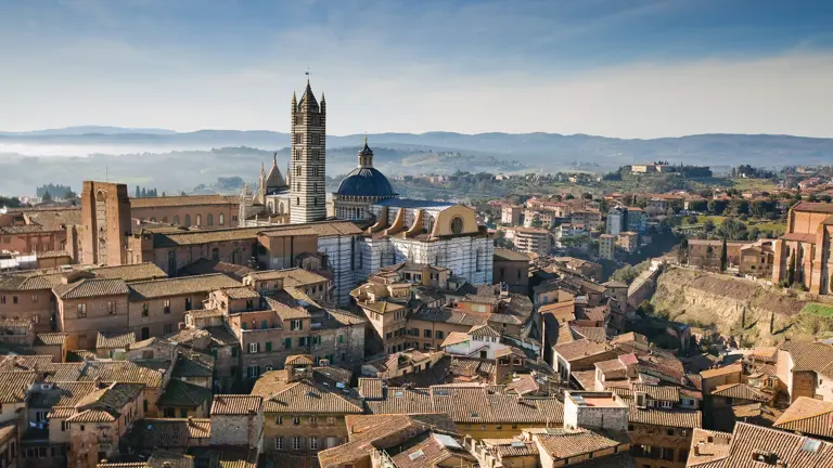The skyline of Siena, the striped marble cathedral and its tall bell tower above orange-tiled roofs, Tuscan hills beyond