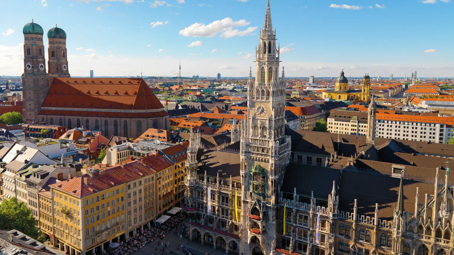 An aerial view over Munich's Marienplatz with the spired Gothic New Town Hall and the green twin-domed towers of the Frauenkirche beyond