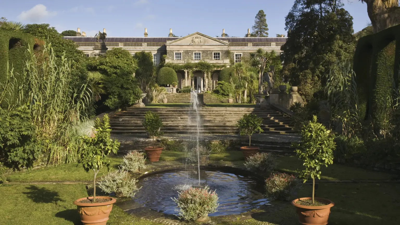 The grey classical front of Mount Stewart House seen across an Italianate garden, a fountain playing in a pool flanked by topiary and statues