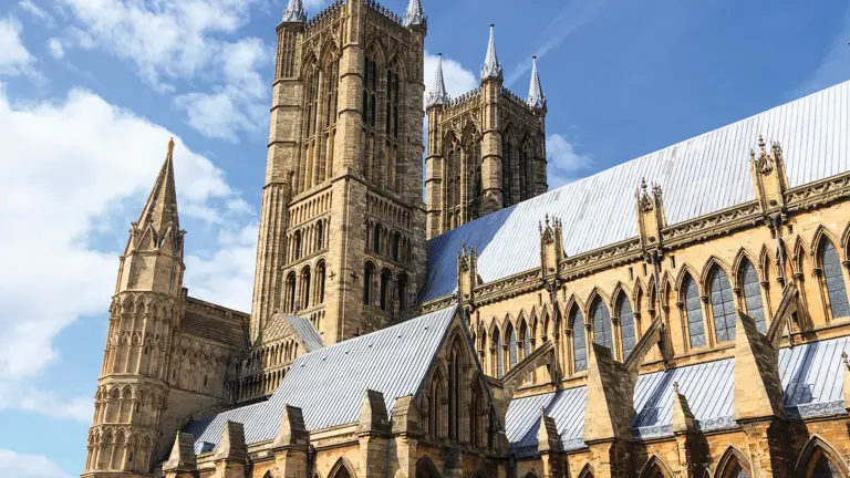 Twin Gothic towers and intricate buttressed nave of Lincoln Cathedral against a bright blue sky, seen from the gardens below the Minster yard