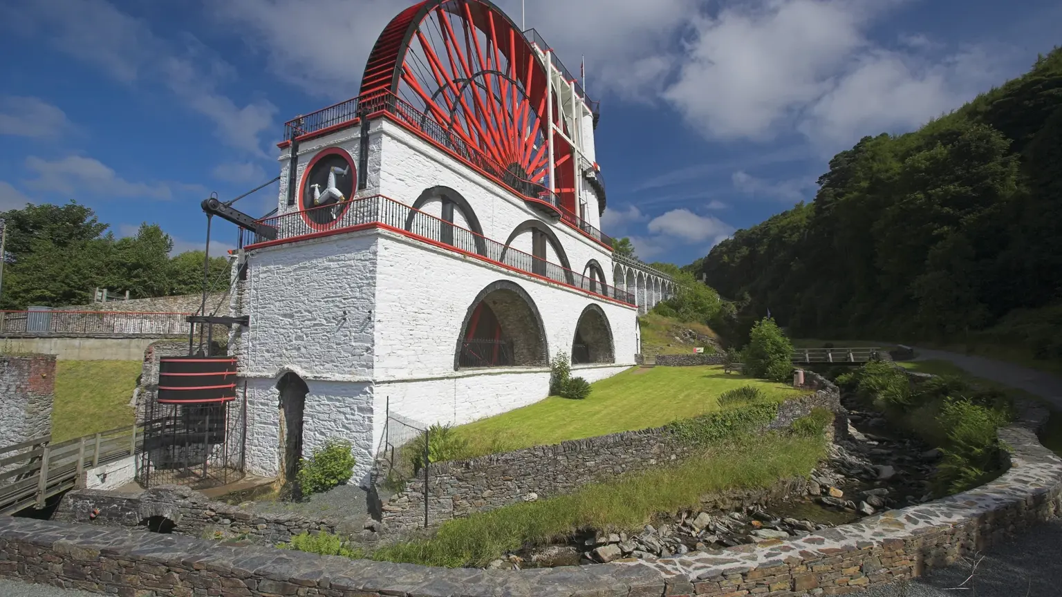 The Great Laxey Wheel, a giant red and white Victorian water wheel set against a hillside at Laxey, with viewing platforms and visitors on the spiral staircase climbing its outer rim