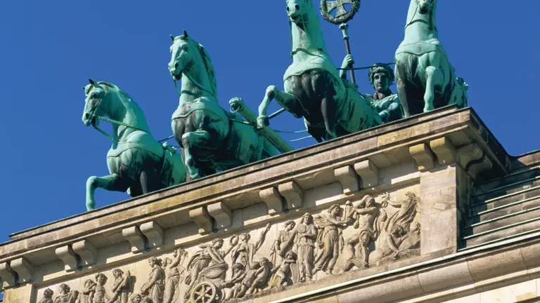 The Quadriga atop the Brandenburg Gate, a bronze chariot drawn by four horses, set against a clear blue sky above the gate's sculpted frieze
