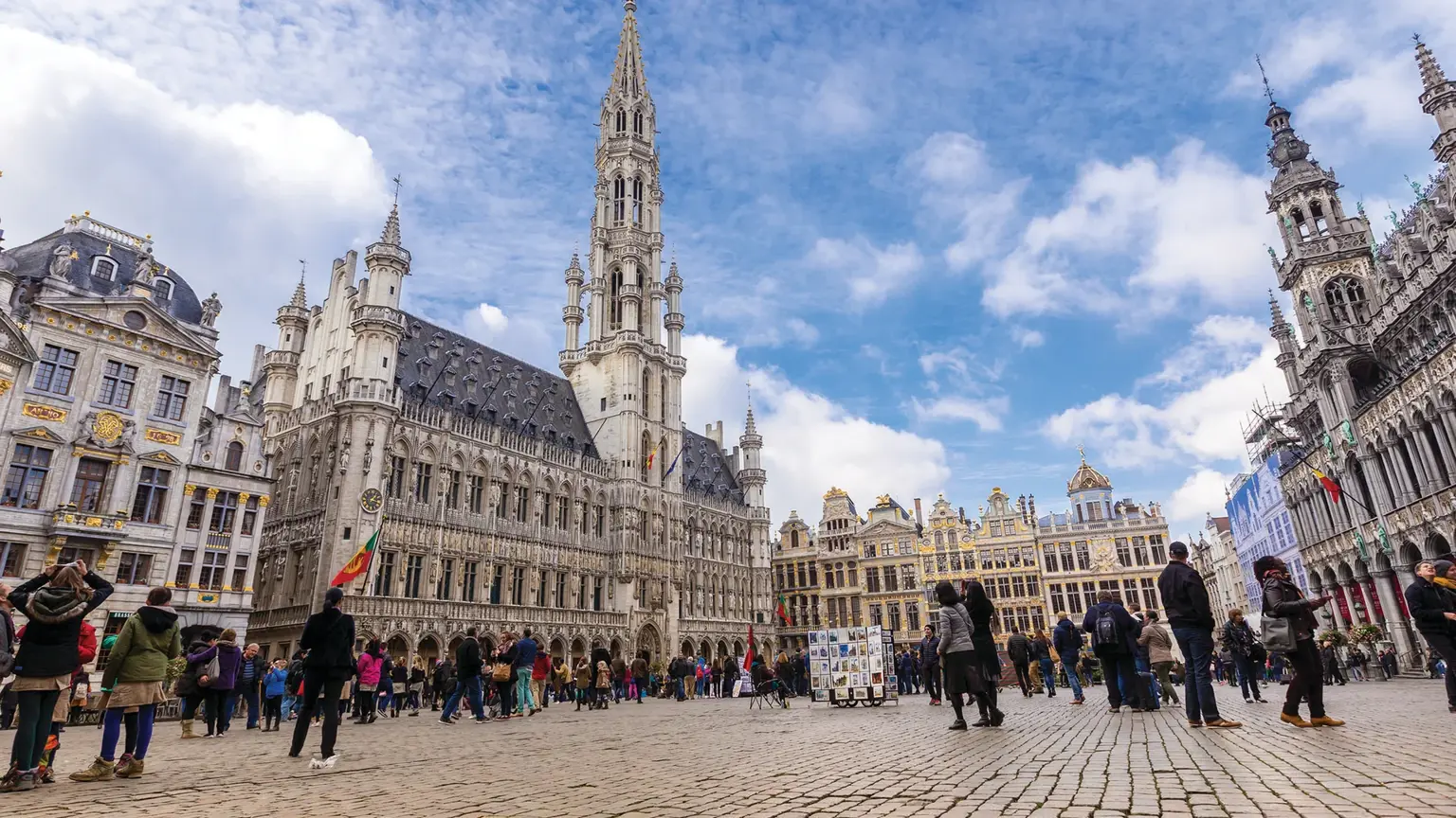 A wide cobbled square dominated by an ornate Gothic town hall with a soaring lacework spire, surrounded by gabled guild houses and crowds.