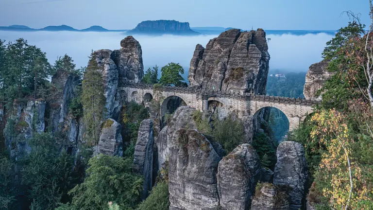 The Bastei stone bridge spanning jagged sandstone pinnacles above a misty valley in Saxon Switzerland, a flat-topped mesa on the horizon