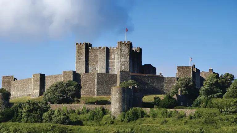 Stone keep and curtain walls of a hilltop medieval castle under a cloudy sky, with green grass in the foreground