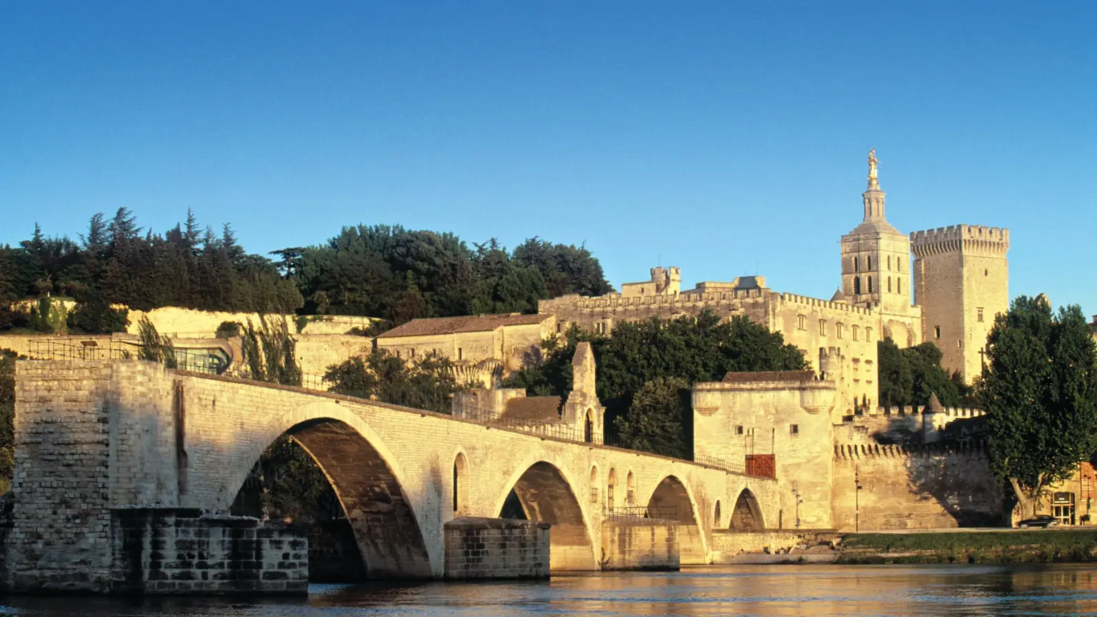 A broken medieval stone bridge reaching into a river below ramparts and a large papal palace at golden hour.