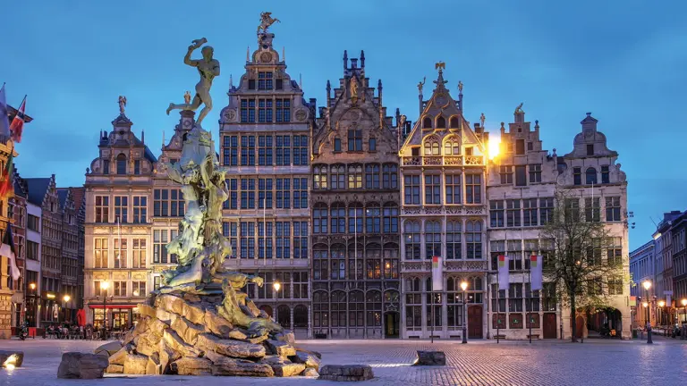 A floodlit row of tall Renaissance guild houses with stepped gables behind a bronze figure fountain on a cobbled square at twilight.