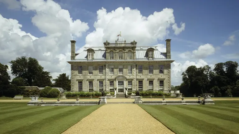 Symmetrical seventeenth century stone mansion with central pediment at the head of a long manicured lawn under a blue sky