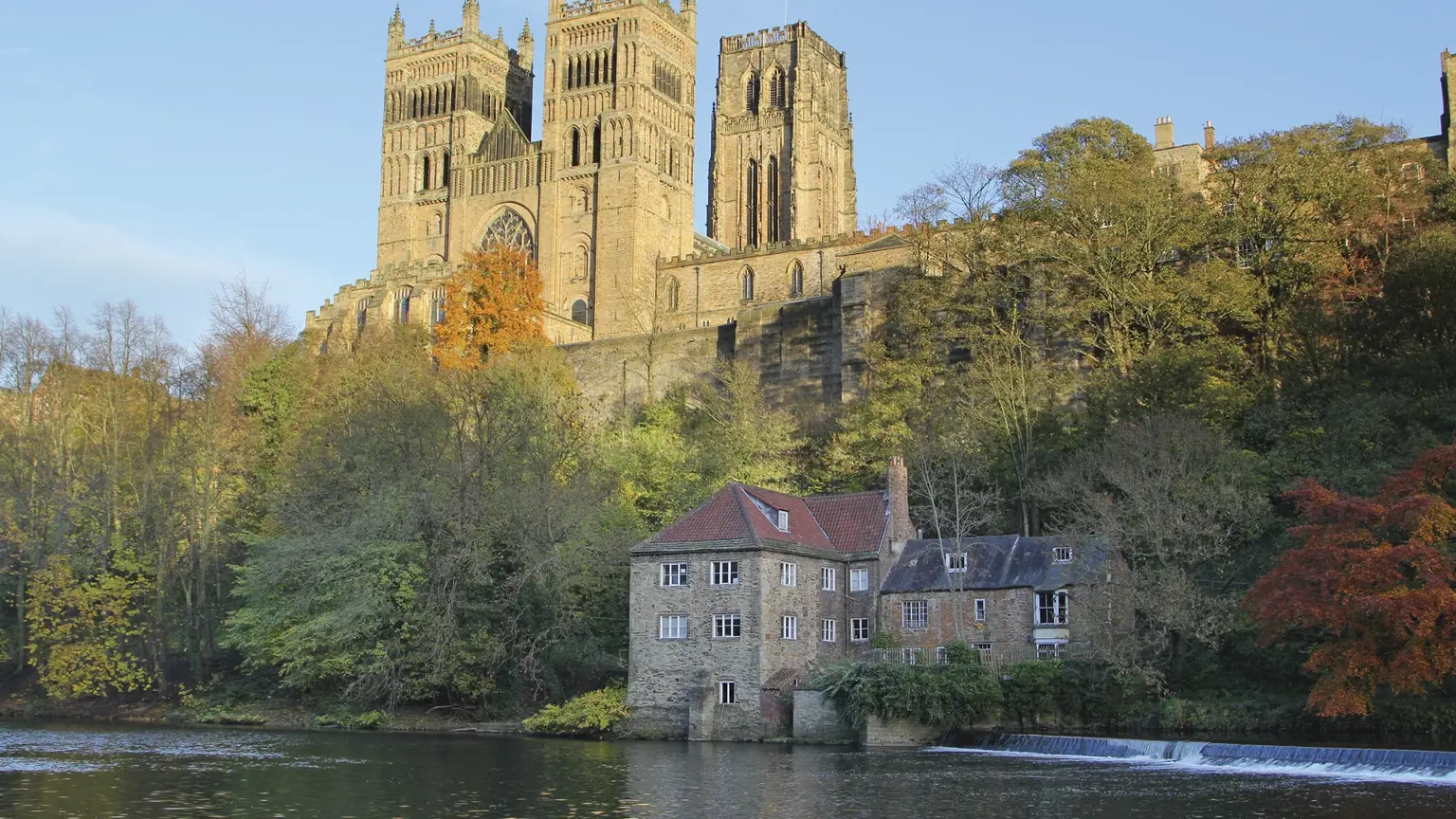 Durham Cathedral's twin Norman towers rising above autumn trees and a riverside fulling mill on the River Wear